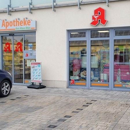 Das Schaufenster einer Apotheke zeigt große Glastüren, ein geparktes Auto und ein Schild mit der Aufschrift "Adler Apotheke" mit Blumen am Eingang.