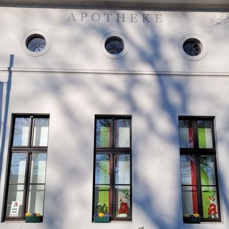 Ein minimalistisches weißes Gebäude mit vier Fenstern, die bunte Dekorationen zeigen, und zwei runden Fenstermotiven darunter. Das Sonnenlicht wirft Schatten an die Wand.